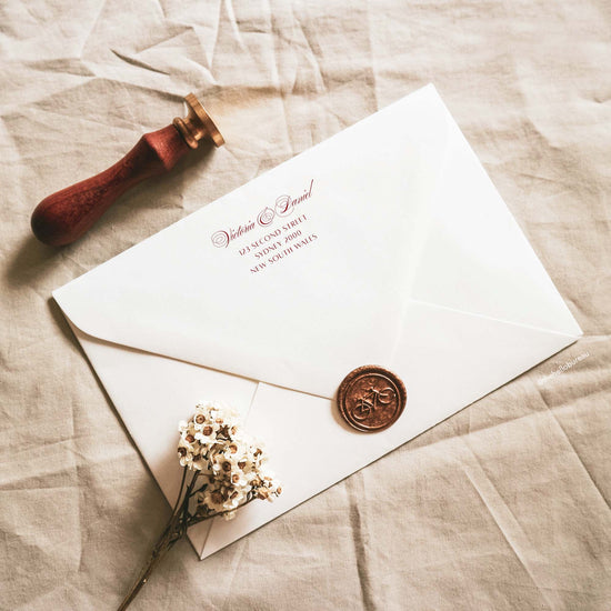 Art Deco wedding envelope return address template printed in dark red on the back flap of a white envelope. A copper wax seal with a bicycle, a wooden stamp and dried white flowers sit on beige linen. The elegant calligraphy text is editable in Canva.