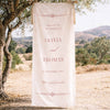 Old money style wedding fabric welcome sign hanging from a tree in a scenic outdoor setting.