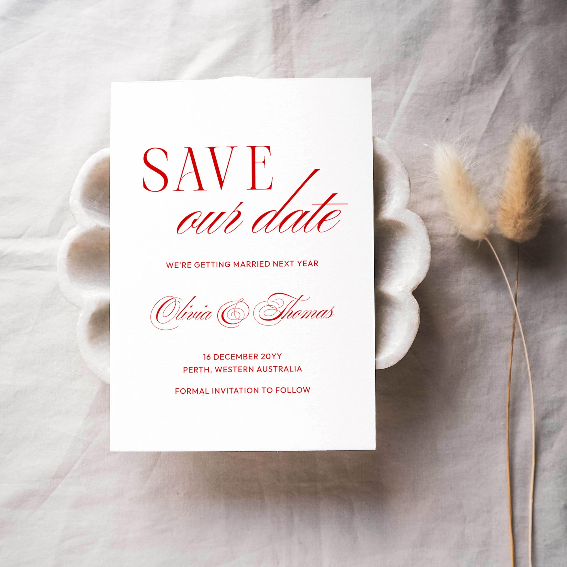 Old-money style save the date card with red text on a white background, placed on a textured surface with dried plants.