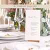 Wedding bar menu card with drink options on a wooden holder sitting on a white table cloth with flowers, plates and wine glasses.