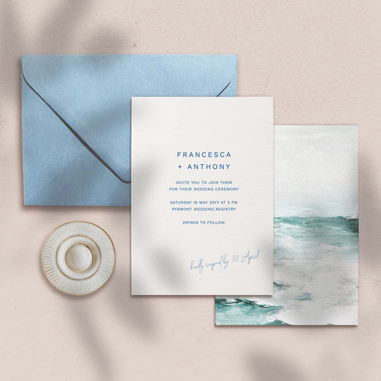 Wedding invitation set with blue envelope and ocean-themed artwork on a beige background.