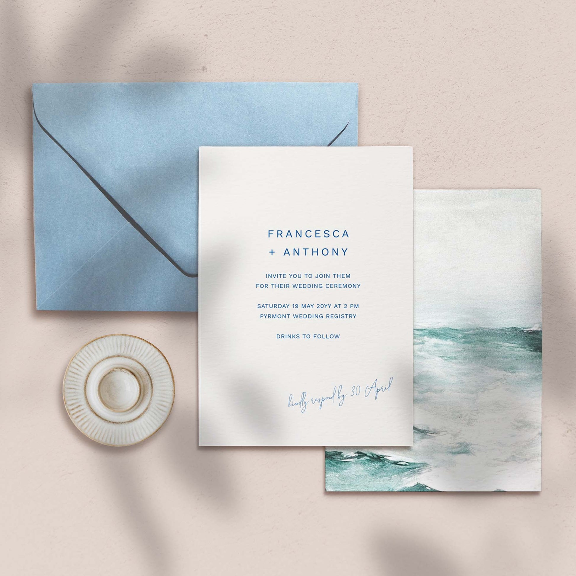 Wedding invitation set with blue envelope and ocean-themed artwork on a beige background.