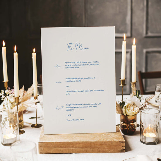 Wedding menu card on a wooden stand with candles and flowers in the background.