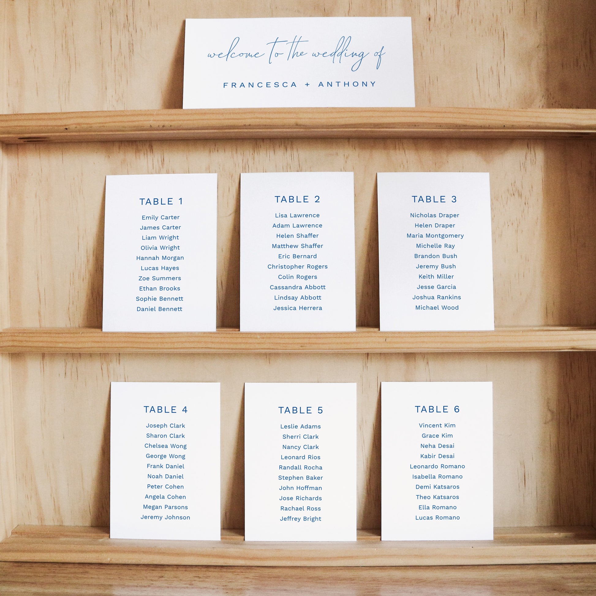 Set of seating cards with names and table numbers on a wooden shelf with a title card at the top.