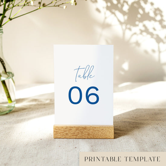A printable table number template displayed on a small wooden block holder on a table with decorative items in the background.