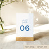 A printable table number template displayed on a small wooden block holder on a table with decorative items in the background.