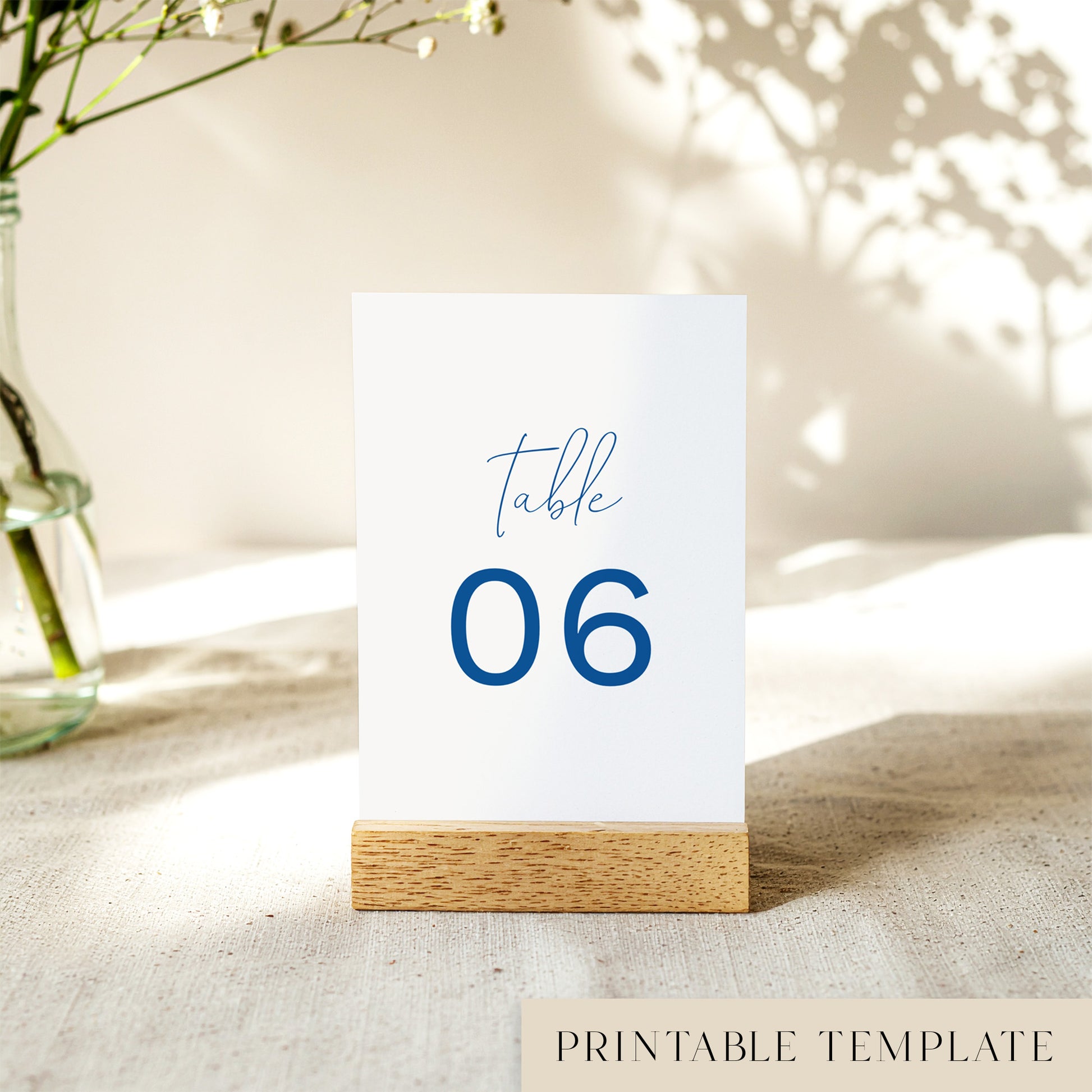 A printable table number template displayed on a small wooden block holder on a table with decorative items in the background.