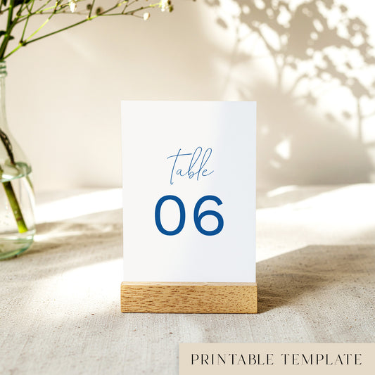 A printable table number template displayed on a small wooden block holder on a table with decorative items in the background.