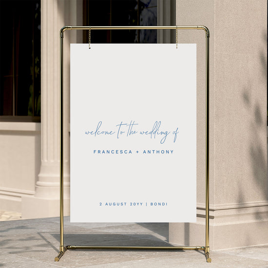 Minimal wedding welcome sign displayed on a gold stand against a neutral wall.