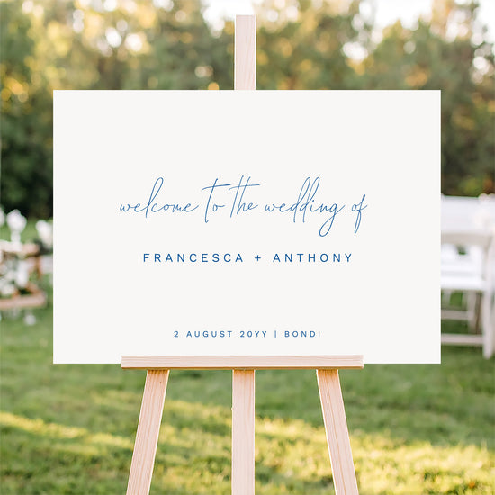Horizontal wedding seating plan sign on an easel with a blurred outdoor ceremony background.