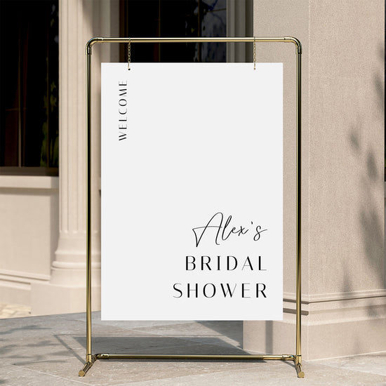 Gold stand with a white welcome sign for 'Alex's Bridal Shower' against a beige wall.
