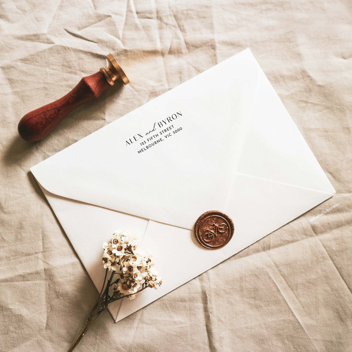 Back of a cream envelope with a printed return address, a copper bicycle wax seal and dried white flowers.