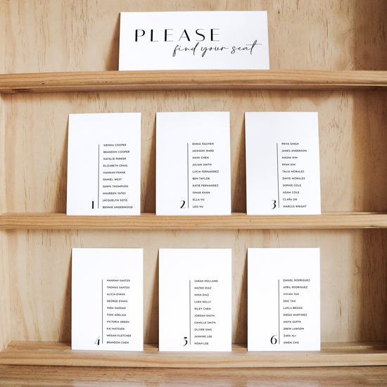 Set of white seating cards with names and table numbers on a wooden shelf with a title card at the top.