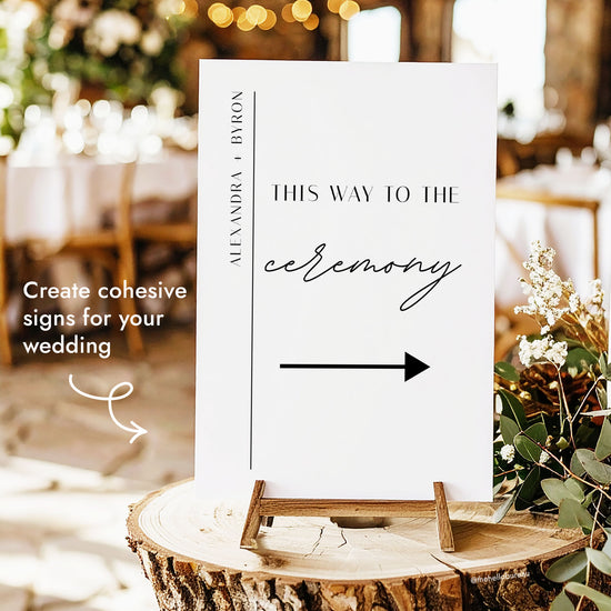 Printable wedding ceremony direction sign template with minimalist line accent, displayed on a wooden stand among greenery and fairy lights.
