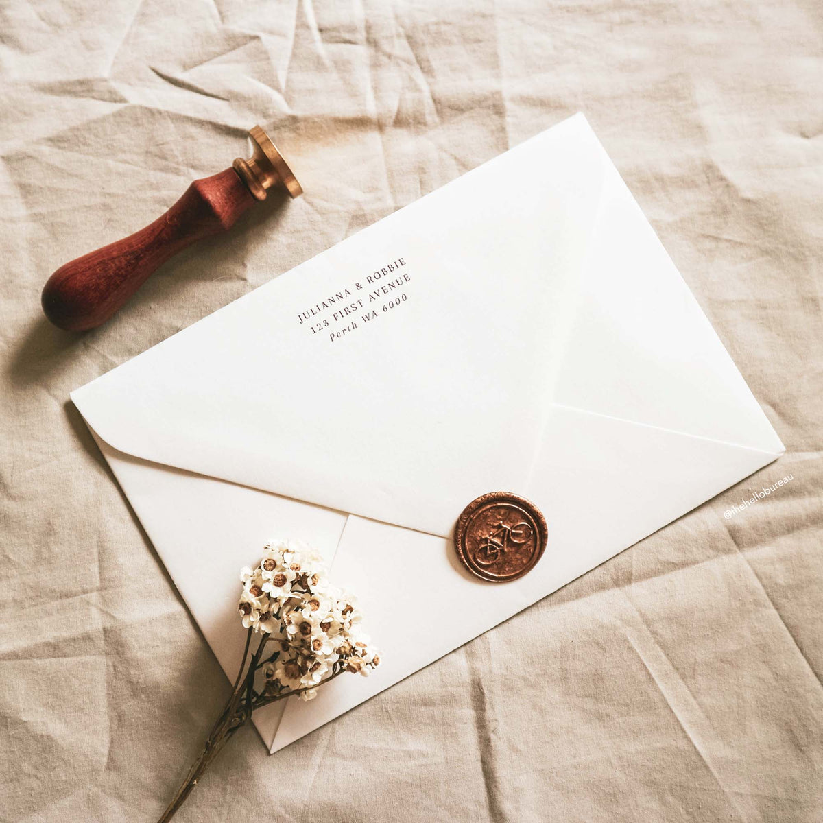 Modern wedding envelope return address template printed on the back flap of a neutral white envelope. A copper wax seal with a bicycle, a wooden stamp and dried white flowers sit on beige linen. The brown text is editable in Canva.