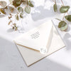 Ivory envelope with an envelope sticker seal featuring a monogram on a light grey surface with eucalyptus leaves.