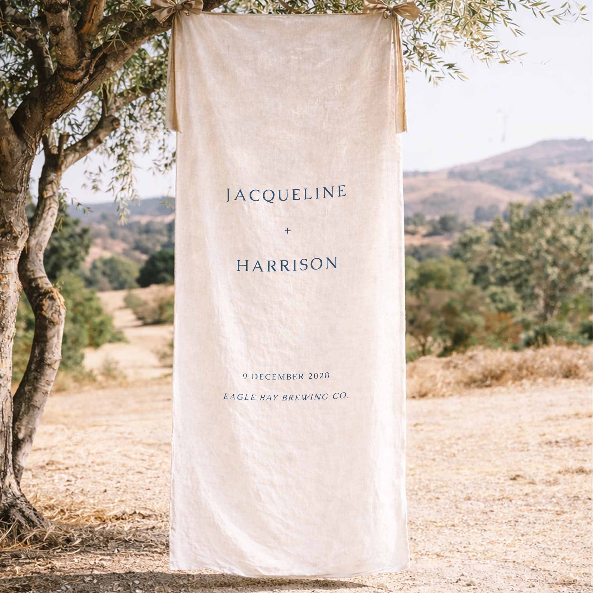 Elegant wedding fabric welcome sign hanging from a tree in a scenic outdoor setting.