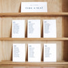 Set of white seating cards with names and table numbers on a wooden shelf with a title card at the top.