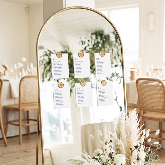 Mirror seating chart display with six modern printable table cards attached with gold wax seals in an elegant light-filled coastal wedding setting.