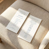 Two DL-sized wedding program cards laid flat on a cushioned chair with sunlight streaming across the linen fabric.