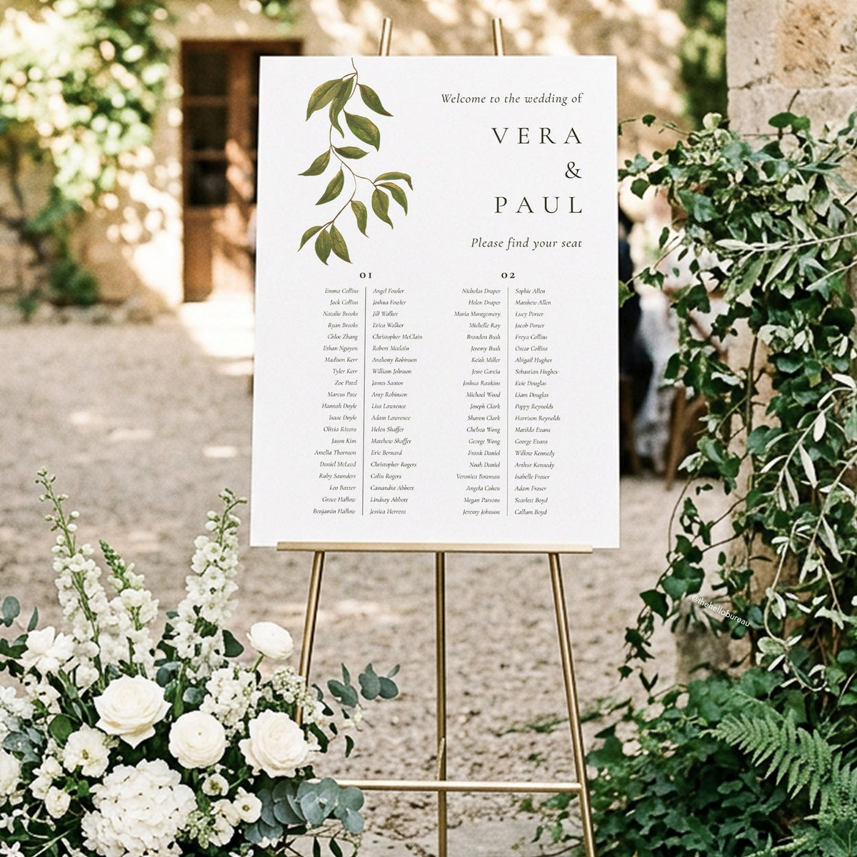 A 2-long-table wedding seating chart on a gold easel, set against a leafy archway and white blooms