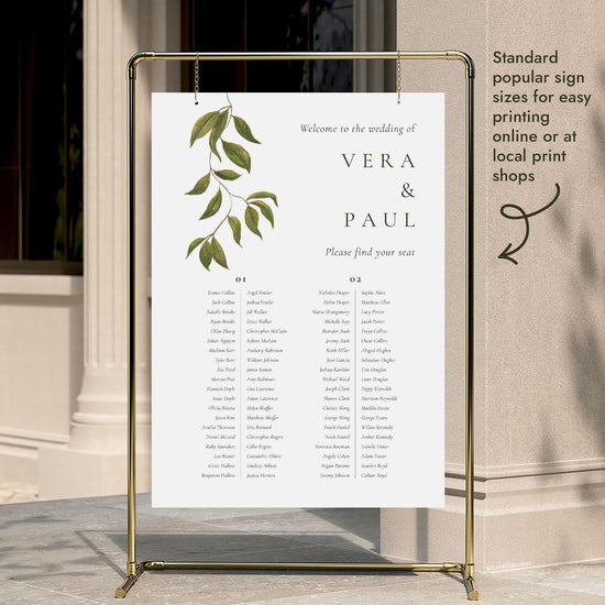 A 2-long-table wedding seating chart template hung on a gold display stand outside a modern city building entrance