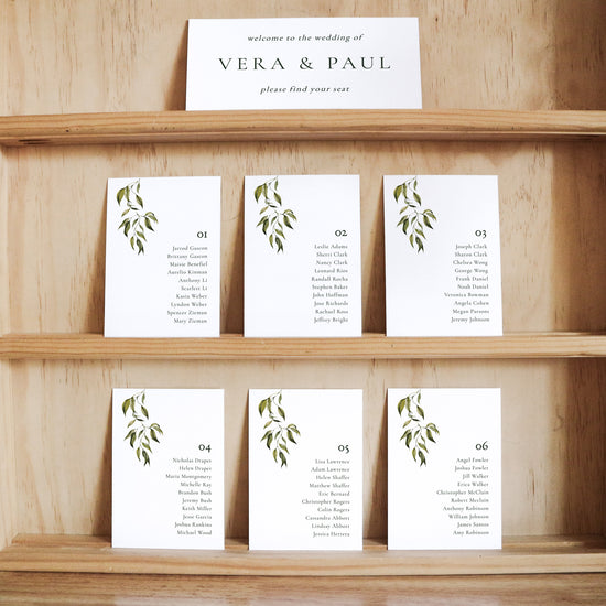 Set of white seating cards with names and table numbers on a wooden shelf with a title card at the top.