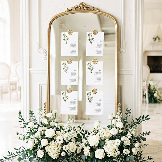 Mirror seating chart display with six minimal leafy printable table cards attached with gold wax seals in an elegant light-filled wedding ballroom setting.
