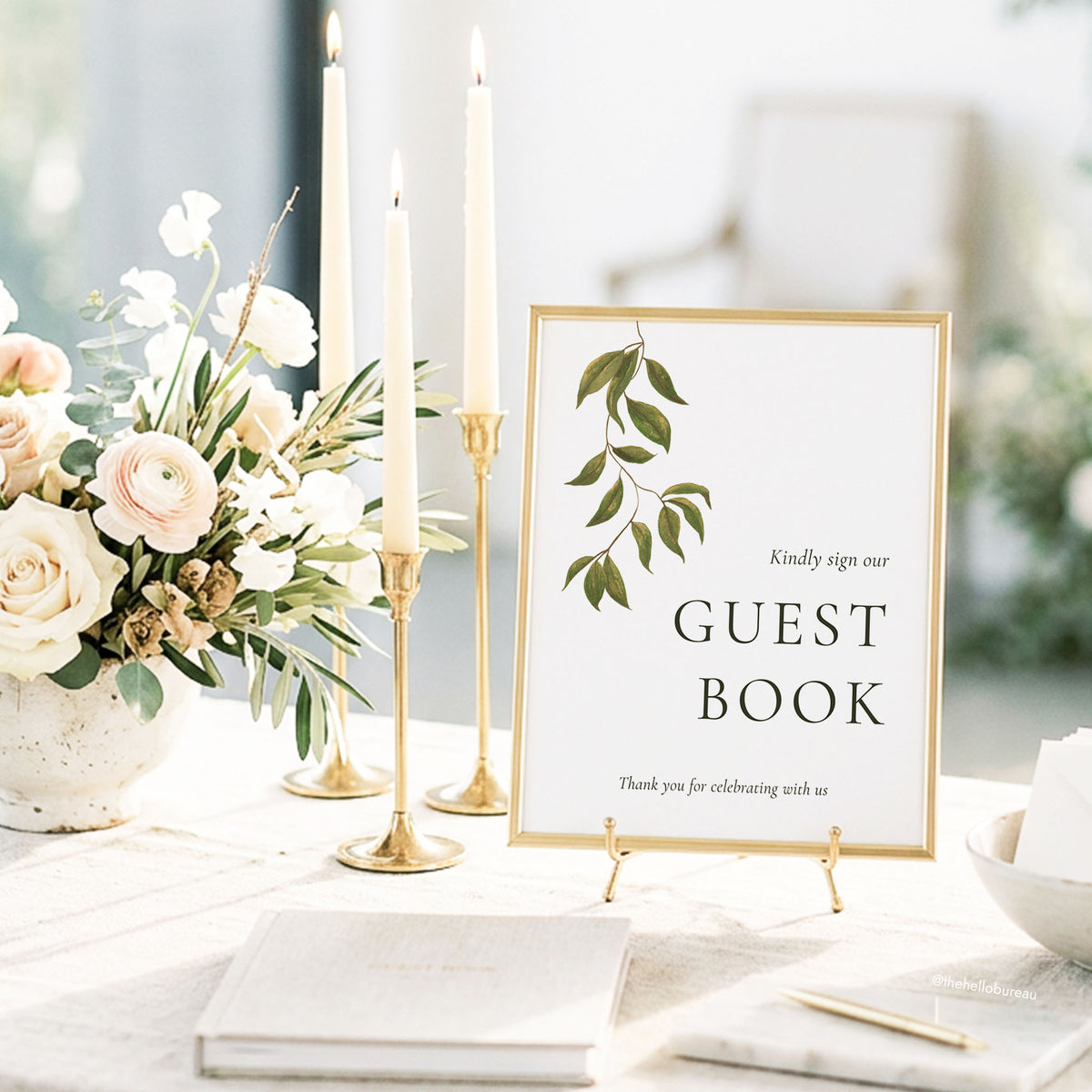 Evergreen guest book sign in a gold frame beside cream roses, ivory candles and a guest book. Editable Canva wedding sign template with a green botanical leaf branch.