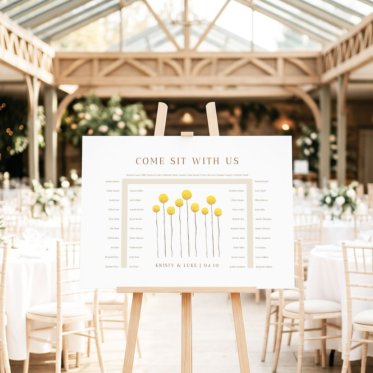 Horseshoe seating chart template featuring yellow billy button wildflowers, shown on a wooden easel inside a sunlit glasshouse wedding venue.