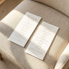 Two DL-sized wedding program cards laid flat on a cushioned chair with sunlight streaming across the linen fabric.