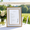 Framed wedding menu with floral arrangement on a table at an outdoors wedding.