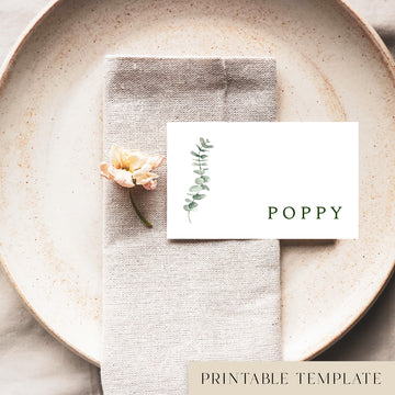 A flat place card with a name printed on it, positioned on a plate with a napkin and a floral decoration.