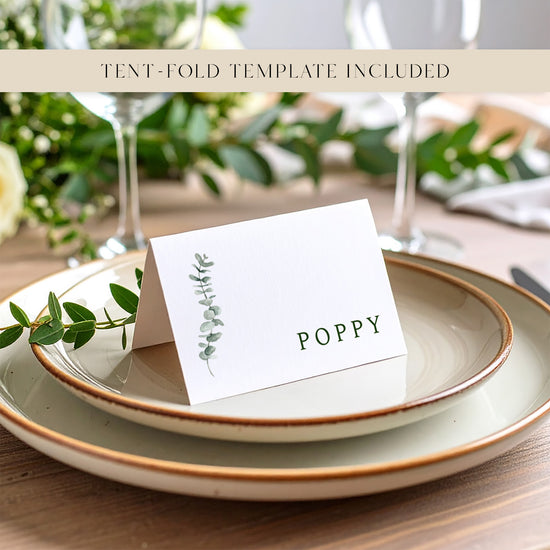 A folded place card with a name printed on it sitting on top of two plates with a eucalyptus leaf for decoration.