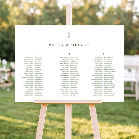 Horizontal wedding seating plan sign on an easel with a blurred outdoor ceremony background.