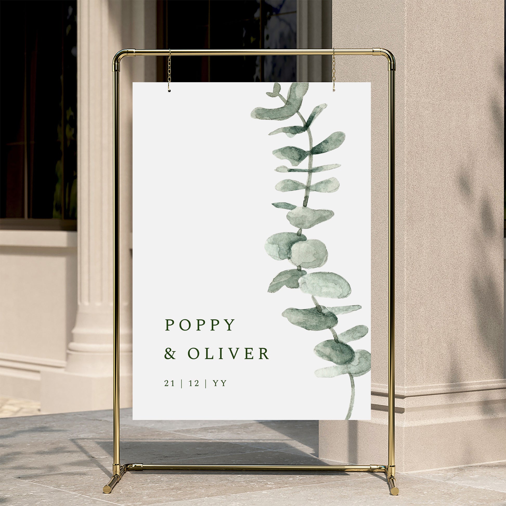 Decorative wedding welcome sign with eucalyptus design and text on a gold stand against a neutral wall.