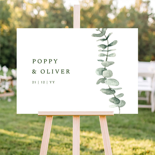 Horizontal wedding welcome sign on an easel with a blurred outdoor ceremony background.