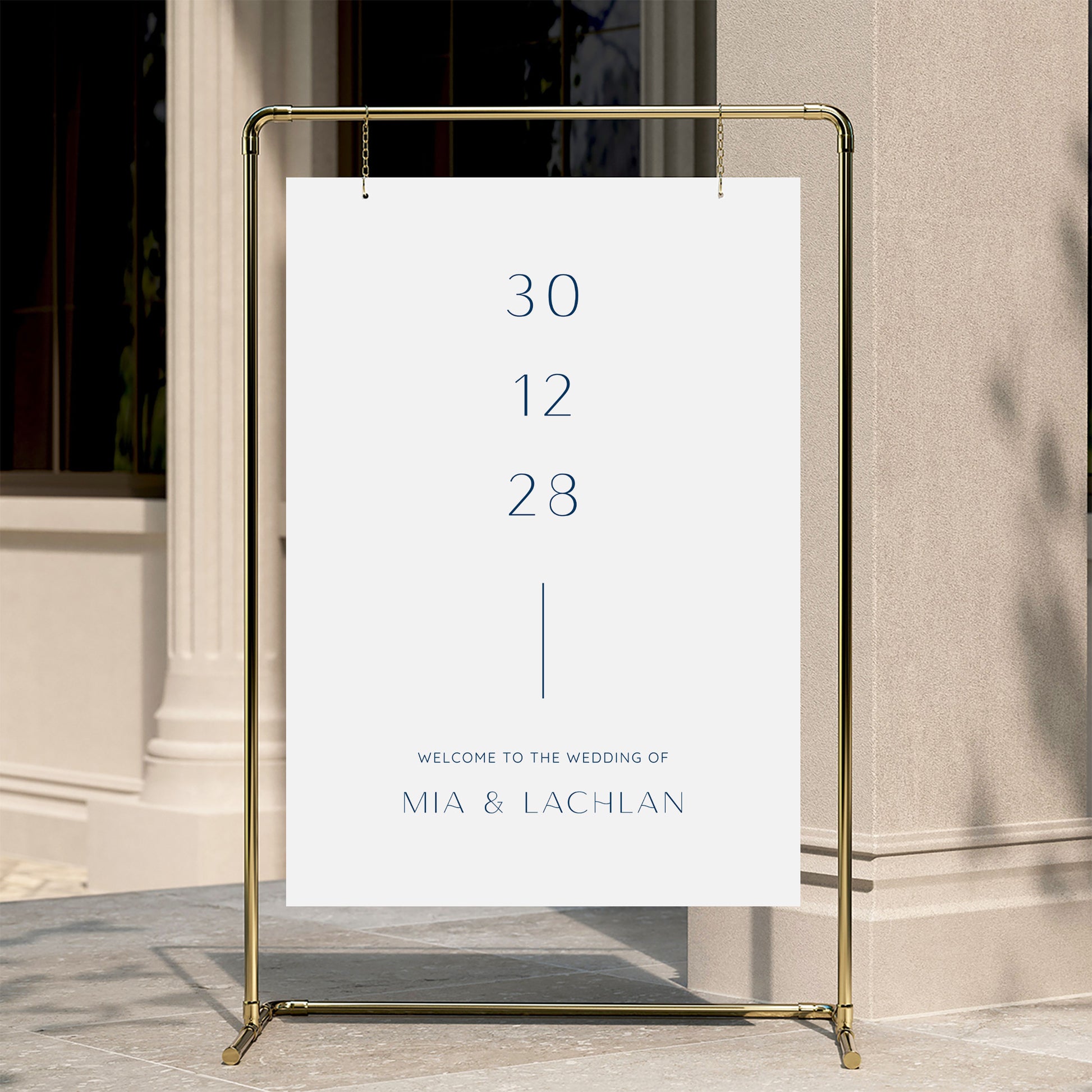 Minimal wedding welcome sign displayed on a gold stand against a neutral wall.