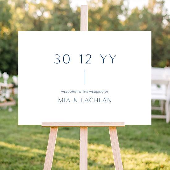 Horizontal wedding welcome sign on an easel with a blurred outdoor ceremony background.