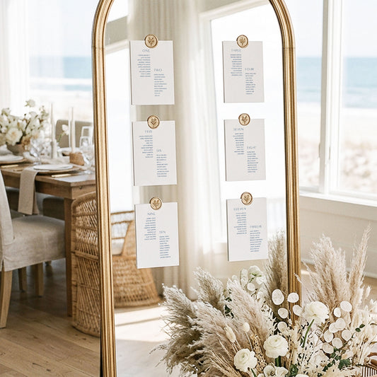 Mirror seating chart display with six minimalistic printable table cards attached with gold wax seals in an elegant light-filled coastal wedding setting.