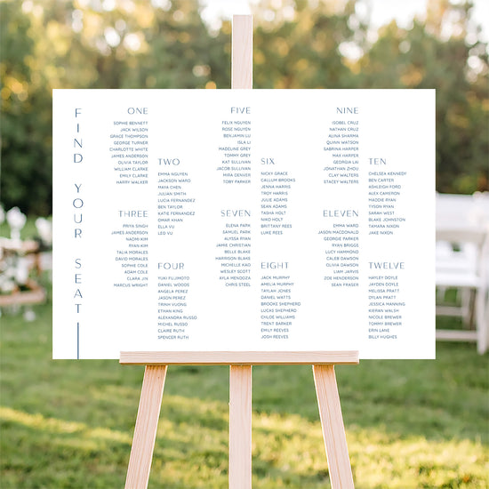Horizontal wedding seating plan sign for long tables on an easel with a blurred outdoor ceremony background.