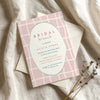 Whimsical pink bridal shower invitation on textured card with envelope on fabric background.