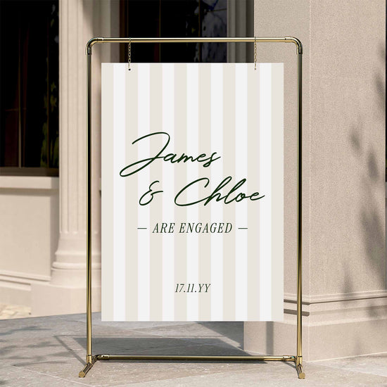 Modern preppy stripe engagement party welcome sign displayed on a gold stand against a neutral wall.