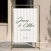 Modern preppy stripe engagement party welcome sign displayed on a gold stand against a neutral wall.