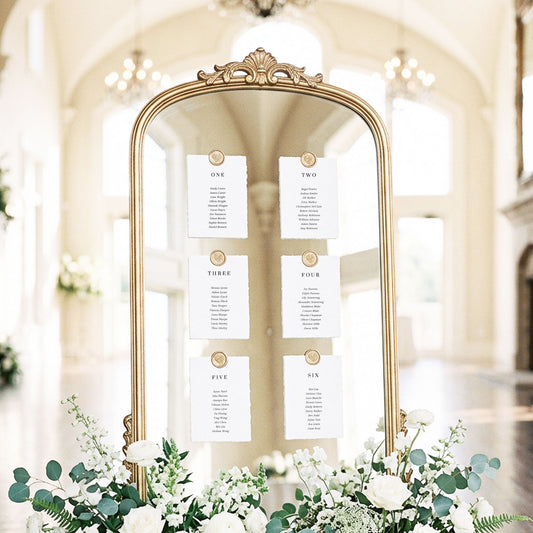 Mirror seating chart display with six minimal printable table cards attached with gold wax seals in an elegant light-filled wedding ballroom setting.