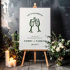 Engagement party sign with champagne glasses on a white board with a wooden easel, surrounded by flowers and candles.