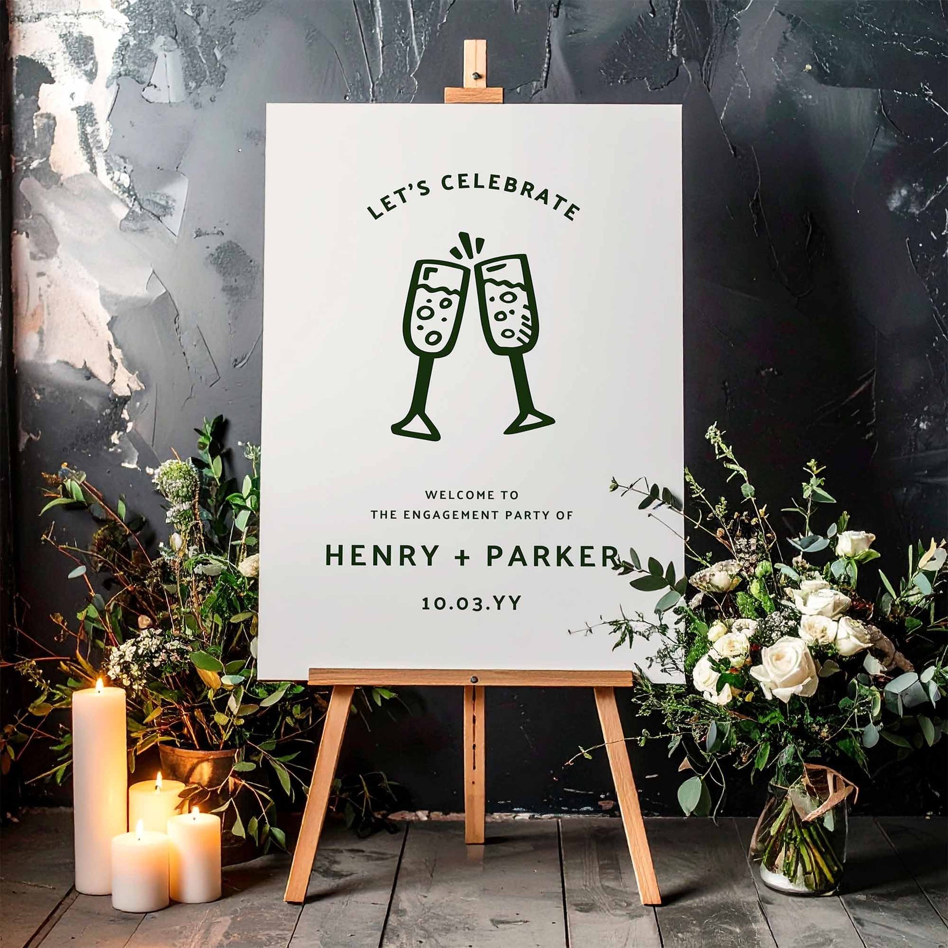 Engagement party sign with champagne glasses on a white board with a wooden easel, surrounded by flowers and candles.