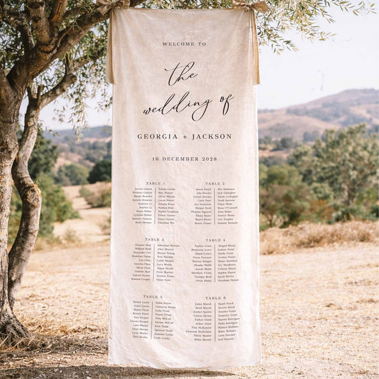 Decorative wedding fabric seating chart sign hanging from a tree in a scenic outdoor setting.