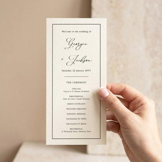 A hand holding a single DL-sized wedding program card in front of a softly lit neutral background.