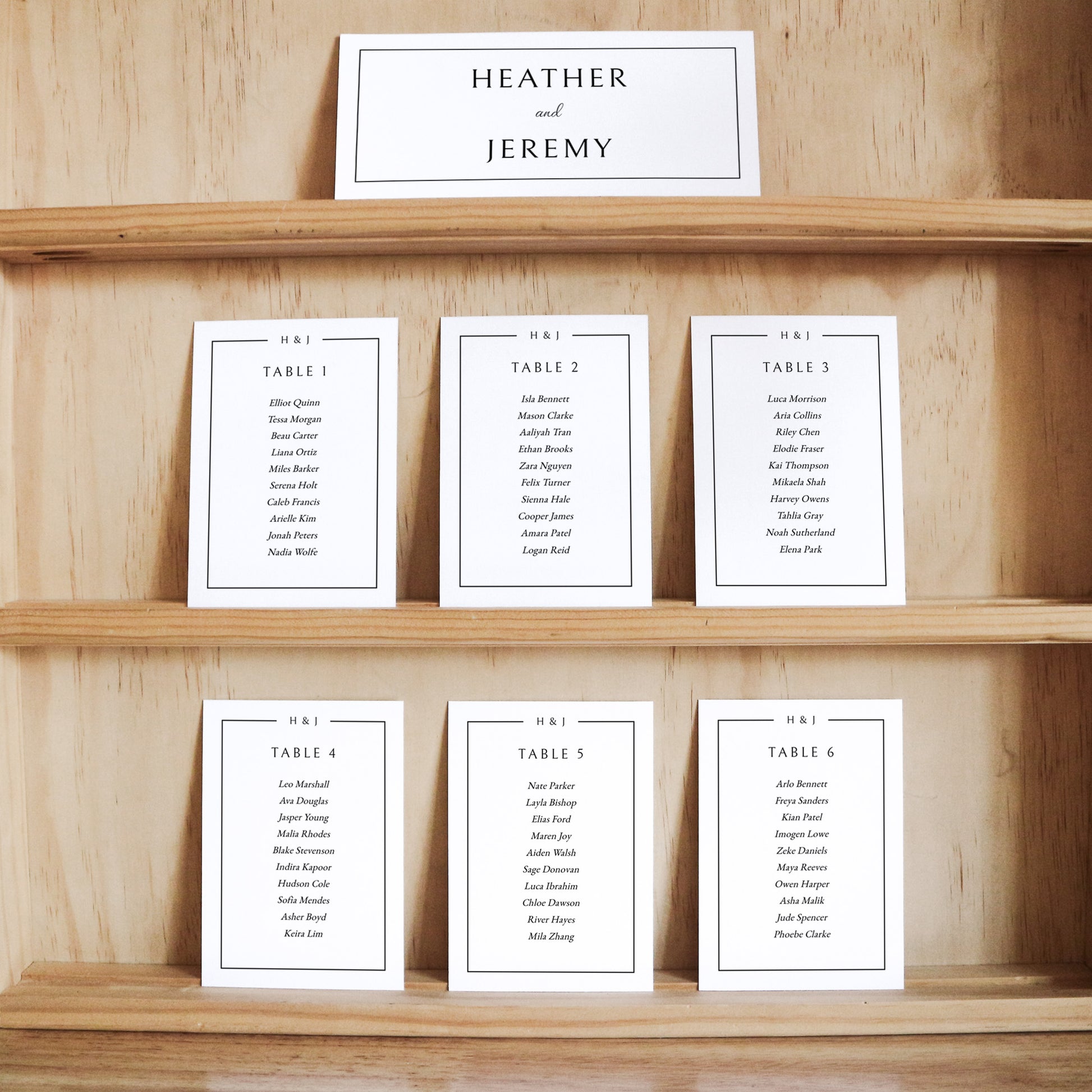 Set of white elegant seating cards with names and table numbers on a wooden shelf with a title card at the top.